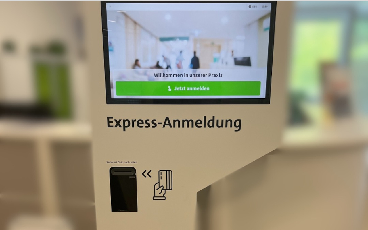 Ein Foto von der "Express-Anmeldung, einer Säule mit Monitor und Fach für den Karteneinschub. Der Tresen steht im Hintergrund in einiger Entfernung. Auf dem Monitor steht: "Herzlich willkommen in unserer Praxis. Jetzt anmelden."